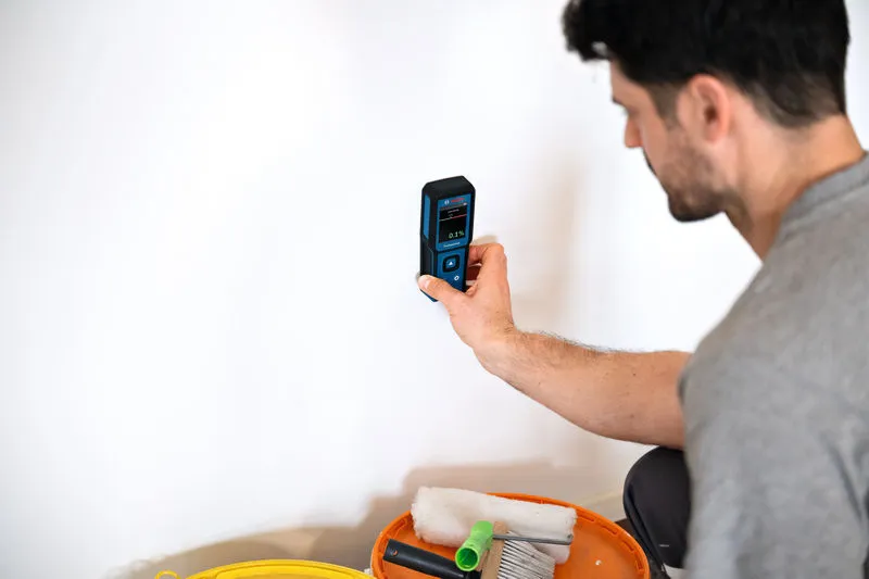 Une personne vérifie l'humidité des murs avec un humidimètre à côté des outils de peinture.