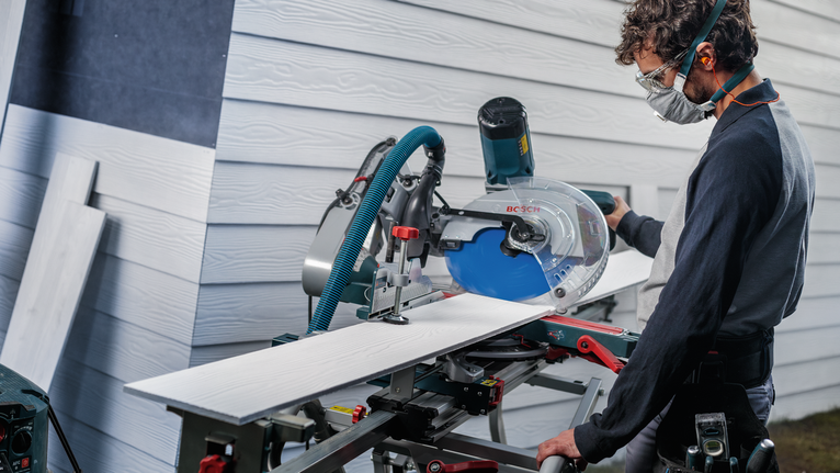 Une personne portant un équipement de sécurité coupe une planche avec une scie circulaire à l'extérieur.
