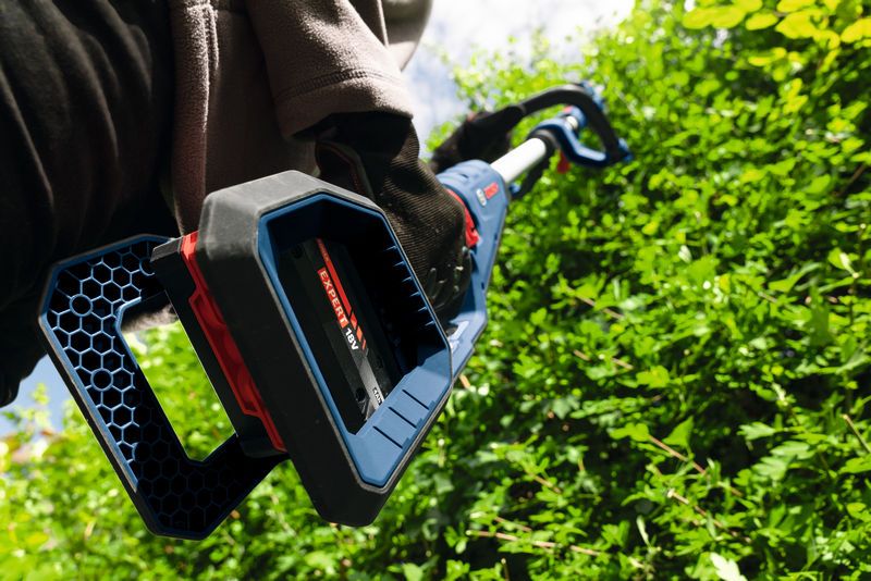 Une personne portant un équipement de sécurité taille des buissons verts denses avec un taille-haie sans fil.