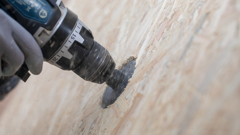 Une personne portant un équipement de sécurité utilise une perceuse électrique avec une scie cloche sur du bois.
