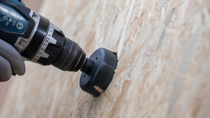 Une personne portant un équipement de sécurité utilise une perceuse sans fil avec une scie cloche pour couper du bois.