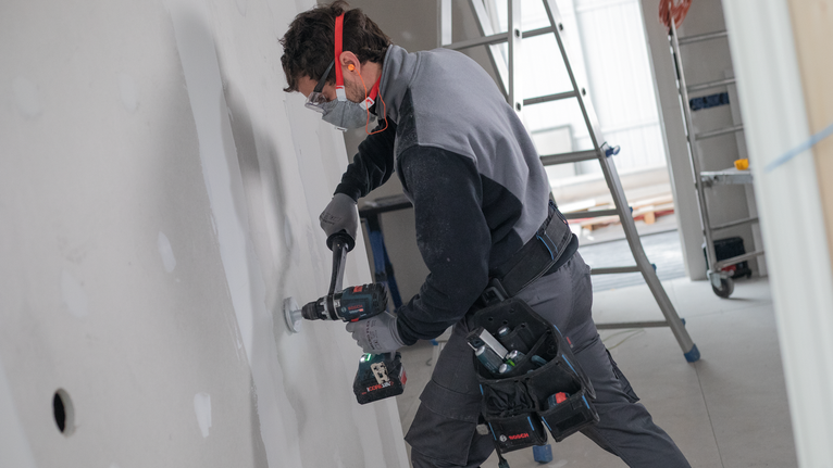 Une personne portant un équipement de sécurité perce un trou dans une cloison sèche dans une zone de construction.
