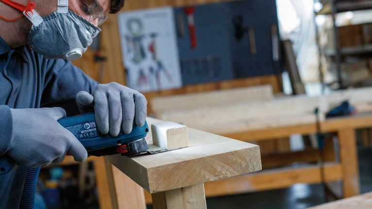 Une personne portant un équipement de sécurité coupe du bois avec un outil électrique dans un atelier.
