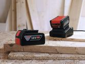 Batterie 18V et chargeur posés sur des planches de bois empilées dans un atelier.