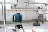 Un niveau laser monté sur trépied projette une ligne rouge pendant qu'une personne perce un mur.