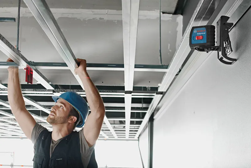 Una persona che indossa dispositivi di sicurezza allinea i telai del soffitto utilizzando uno strumento di livellamento laser.