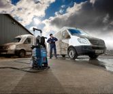 Una persona che indossa un equipaggiamento di sicurezza utilizza un'idropulitrice per pulire un furgone sporco all'aperto.