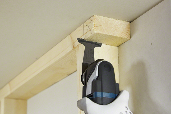 Una persona che indossa dispositivi di protezione individuale utilizza un utensile multifunzione per tagliare il legno in un angolo del soffitto.