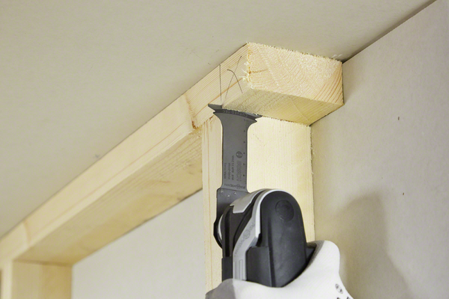 Una persona che indossa dispositivi di sicurezza taglia il legno con un utensile multiuso vicino a un angolo del soffitto.