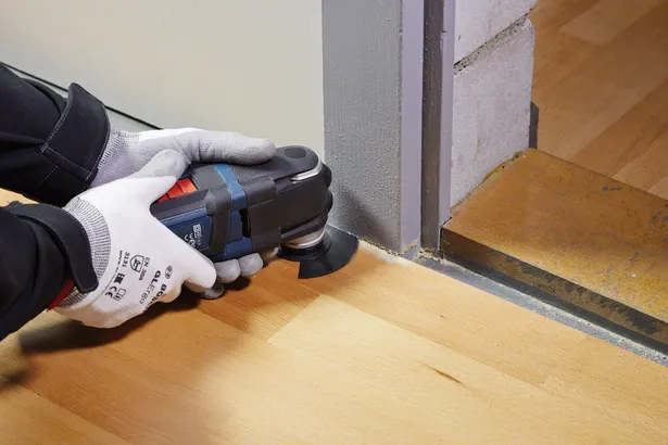 Una persona che indossa dispositivi di sicurezza rifinisce il pavimento in legno con un utensile multiuso vicino allo stipite di una porta.