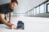 Una persona traccia una linea sul pavimento utilizzando una livella laser in una stanza luminosa e vuota.