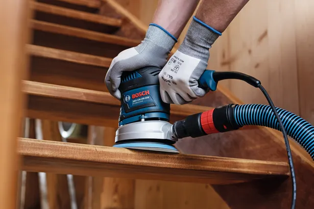 Un lavoratore che indossa dispositivi di sicurezza leviga un gradino di una scala in legno utilizzando una levigatrice elettrica collegata.