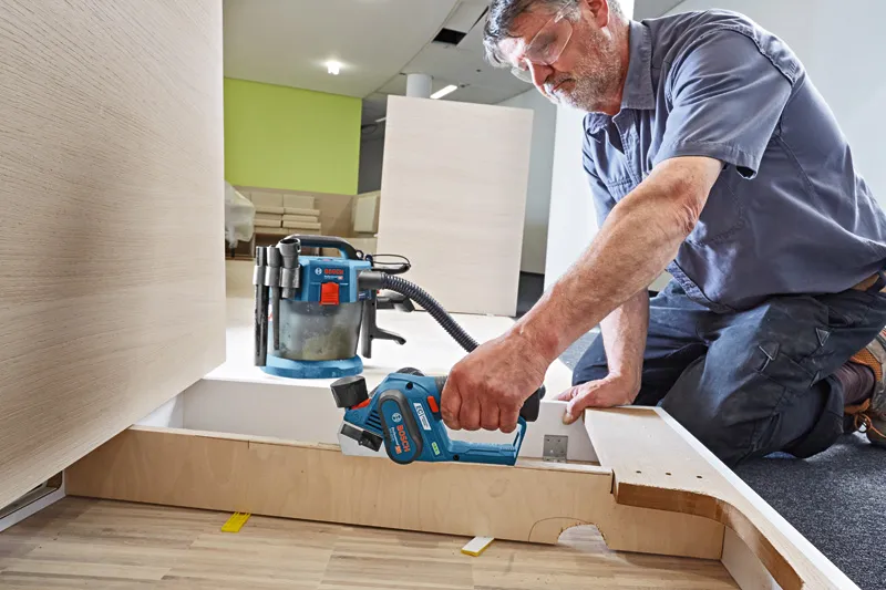 Una persona che indossa dispositivi di sicurezza utilizza una pialla a batteria per levigare pannelli di legno.