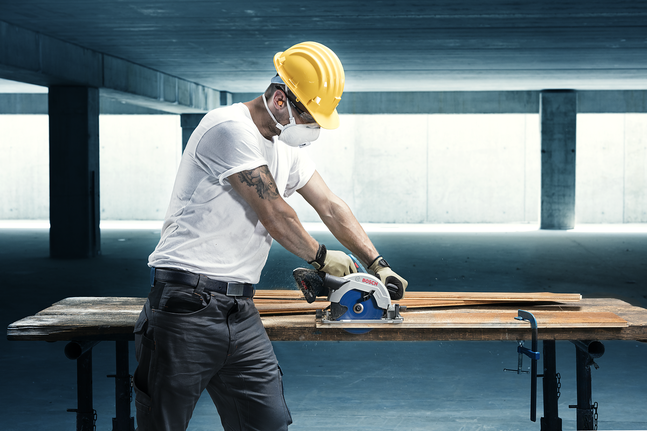 Una persona che indossa dispositivi di protezione individuale utilizza una sega circolare per tagliare il legno su un banco da lavoro.