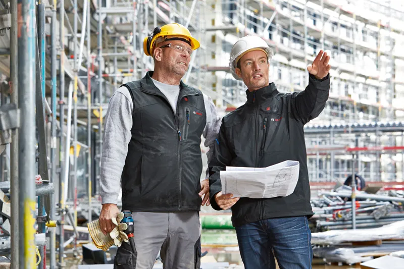 Due operai che indossano dispositivi di sicurezza discutono i progetti di un edificio in un cantiere.