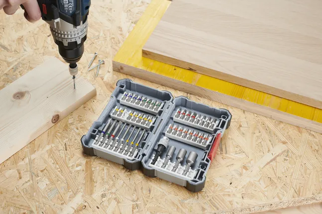 Una persona avvita una vite nel legno con un trapano, accanto a un set di punte aperto.