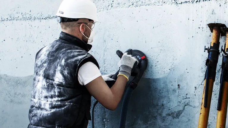 Una persona che indossa un equipaggiamento di sicurezza lucida un muro di cemento con un elettroutensile.