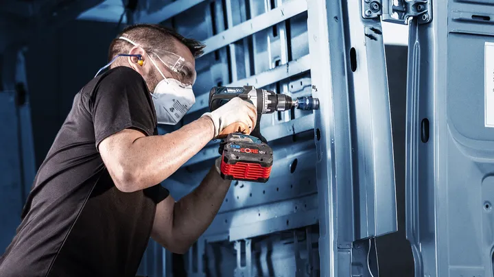 Persona che indossa dispositivi di sicurezza mentre utilizza un trapano a batteria per fissare il metallo all'interno di un veicolo.