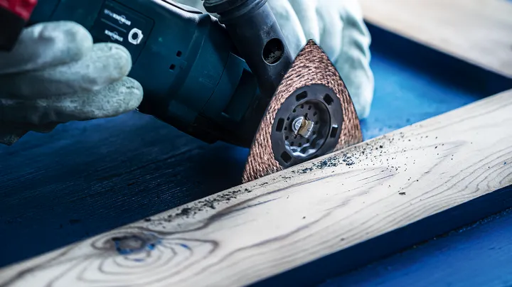 Una persona che indossa l'equipaggiamento di sicurezza leviga una tavola di legno con un utensile multiuso.