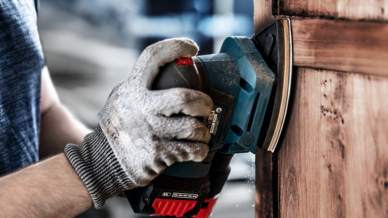 Persona che indossa dispositivi di sicurezza mentre leviga una porta di legno con una levigatrice elettrica.