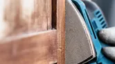 Una persona che indossa dispositivi di sicurezza carteggia una porta di legno con una levigatrice di precisione.