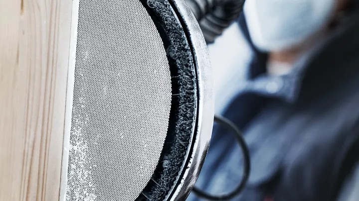 Persona che indossa dispositivi di sicurezza mentre leviga una superficie di legno con un utensile elettrico.