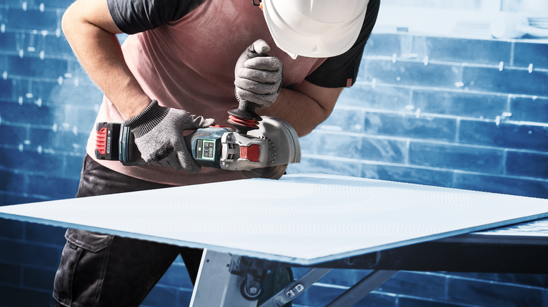 Una persona che indossa dispositivi di sicurezza macina un foglio di materiale su un banco da lavoro.