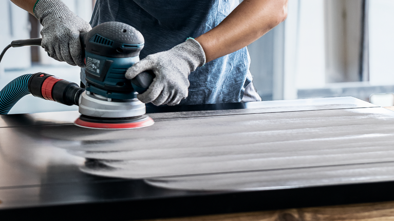 Una persona che indossa dispositivi di sicurezza leviga una superficie di legno con un utensile elettrico.