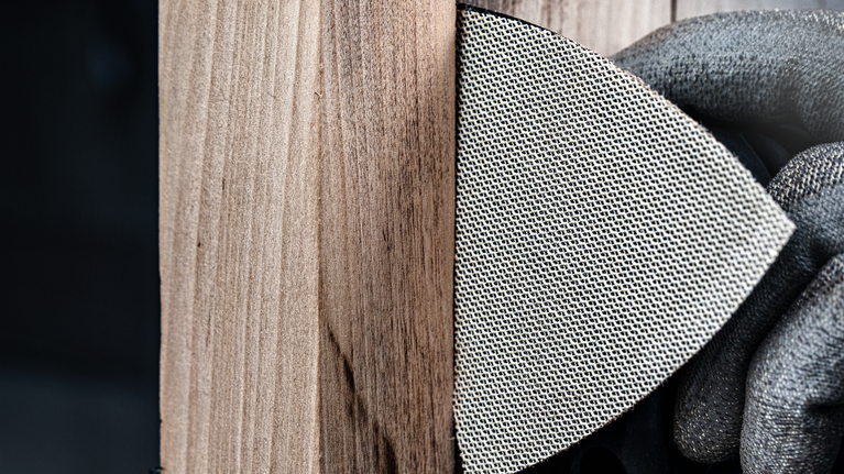 Una persona che indossa dispositivi di sicurezza leviga una superficie di legno con un tampone abrasivo in rete.