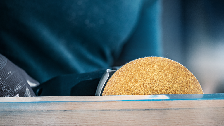 Una persona che indossa dispositivi di sicurezza leviga il legno con una levigatrice elettrica portatile.