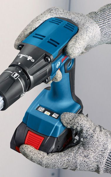 Una persona che indossa dispositivi di protezione individuale tiene un trapano a batteria blu contro un muro.