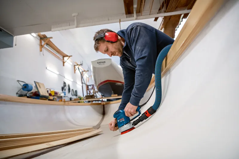 Una persona che indossa l'equipaggiamento di sicurezza carteggia l'interno dello scafo di una barca in legno.