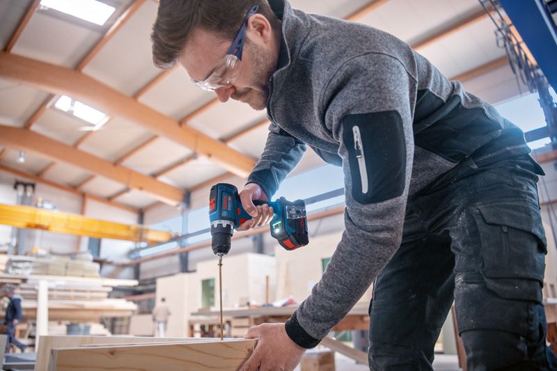 Una persona che indossa dispositivi di sicurezza fora il legno con un utensile elettrico a batteria.