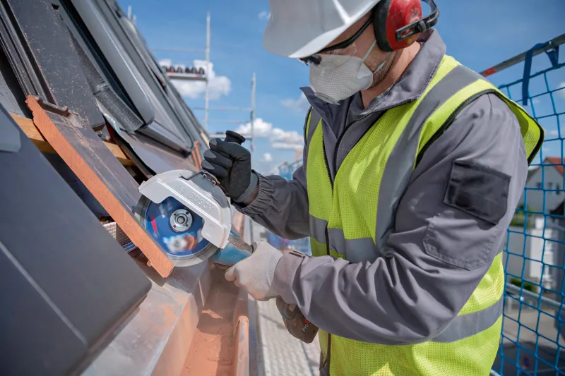 Una persona che indossa dispositivi di sicurezza utilizza una smerigliatrice angolare a batteria per tagliare le tegole.