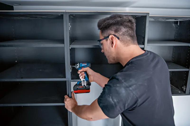 Una persona che indossa dispositivi di sicurezza monta le scaffalature con un trapano o un avvitatore a batteria.