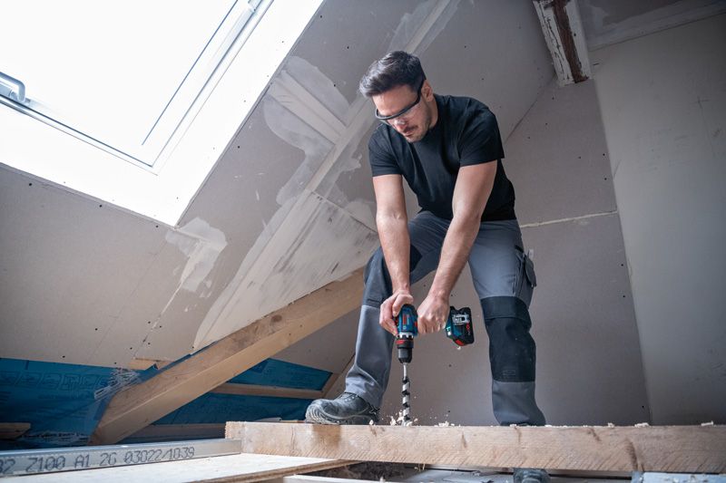 Una persona che indossa un equipaggiamento di sicurezza fora una trave di legno in una soffitta parzialmente rifinita.