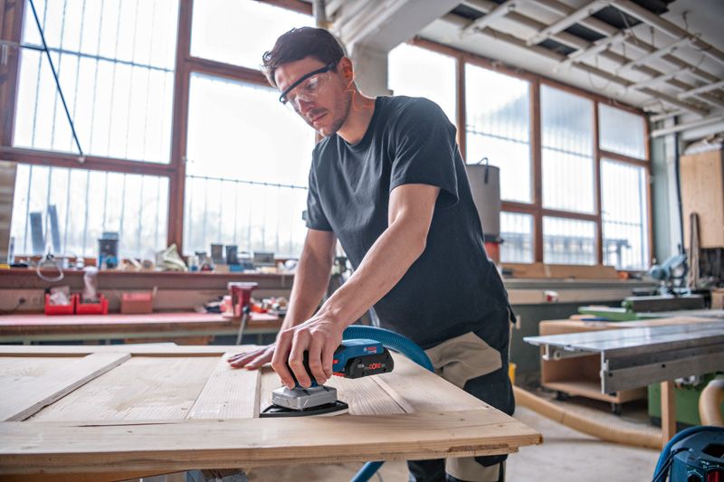 Una persona che indossa dispositivi di sicurezza leviga una porta di legno con una levigatrice orbitale a batteria.