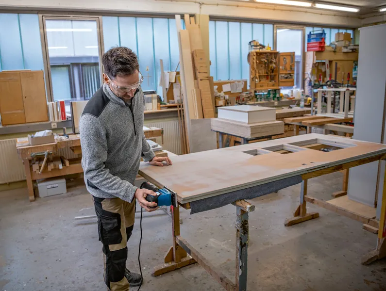 Una persona che indossa dispositivi di sicurezza leviga una porta di legno utilizzando una levigatrice orbitale.