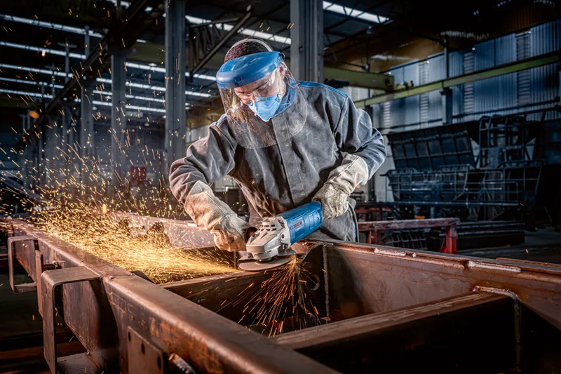 Una persona che indossa un equipaggiamento di sicurezza macina il metallo con una grande smerigliatrice angolare, producendo scintille.