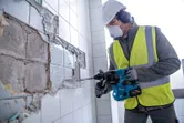 Una persona che indossa dispositivi di sicurezza rimuove le piastrelle da un muro utilizzando un martello perforatore.