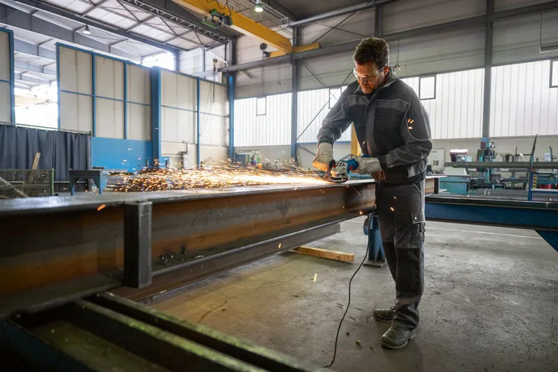 Una persona che indossa un equipaggiamento di sicurezza macina una trave di acciaio, creando scintille luminose.