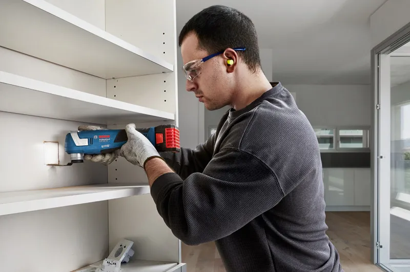 Una persona che indossa un equipaggiamento di sicurezza taglia il pannello di uno scaffale con un utensile multiuso.