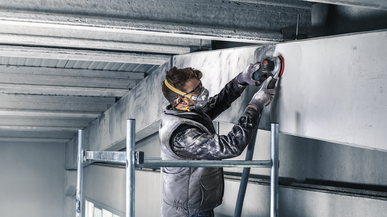 Una persona che indossa un equipaggiamento di sicurezza leviga una trave di cemento utilizzando un utensile elettrico.