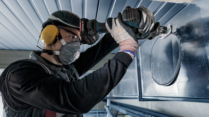 Una persona che indossa un equipaggiamento di sicurezza pratica un foro circolare in una conduttura metallica con un elettroutensile.