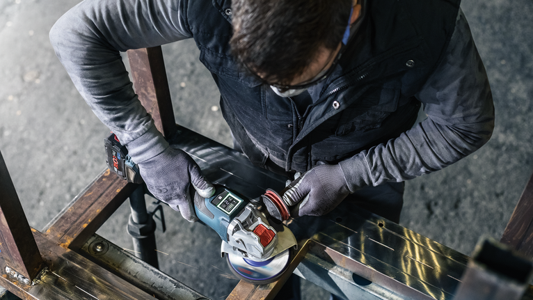 Una persona che indossa un equipaggiamento di sicurezza leviga una trave metallica con una smerigliatrice angolare.