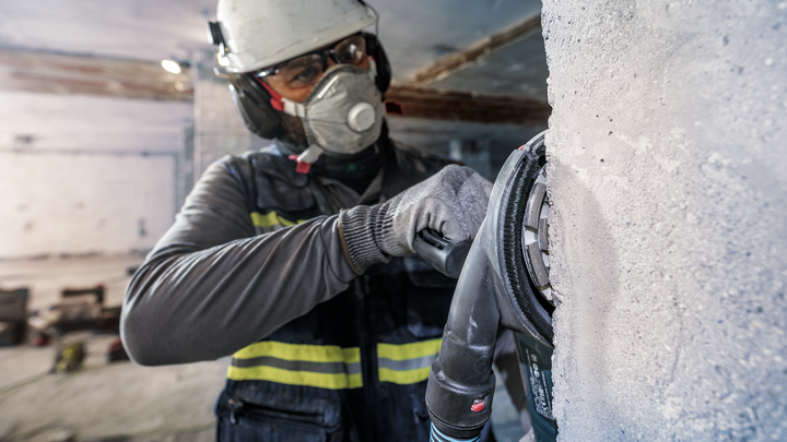 Una persona che indossa dispositivi di sicurezza leviga un muro di cemento con un utensile elettrico.