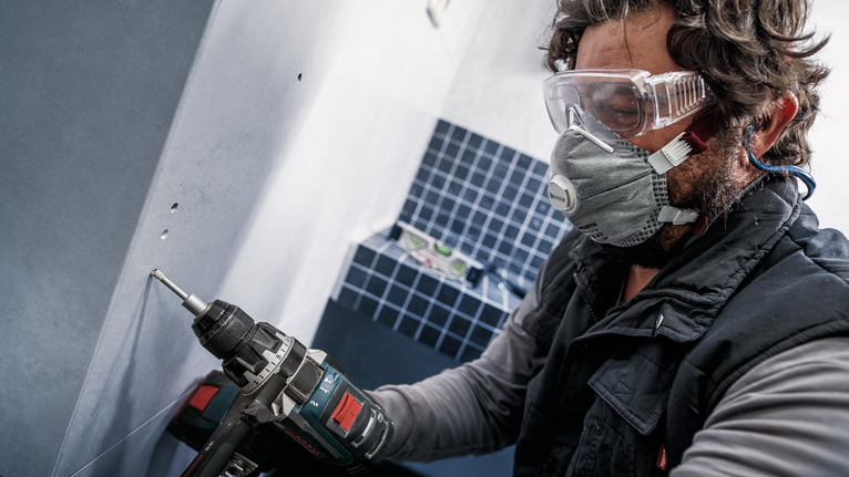 Una persona che indossa dispositivi di sicurezza fora un muro in un'area di lavoro piastrellata.