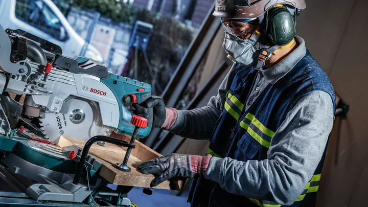 Una persona che indossa un equipaggiamento di sicurezza utilizza una sega troncatrice per tagliare una tavola di legno.