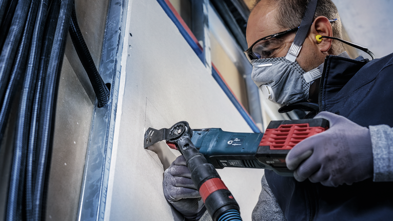 Una persona che indossa dispositivi di sicurezza taglia il cartongesso con un utensile multifunzione elettrico.