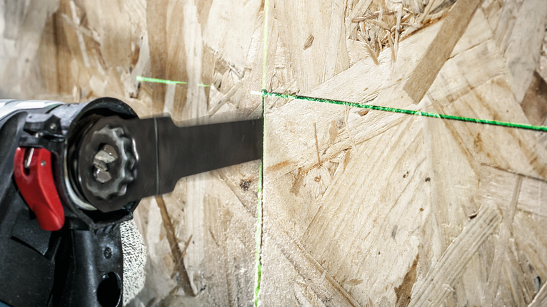 Una persona che indossa un equipaggiamento di sicurezza utilizza una sega alternativa per tagliare il legno lungo una linea guida verde.